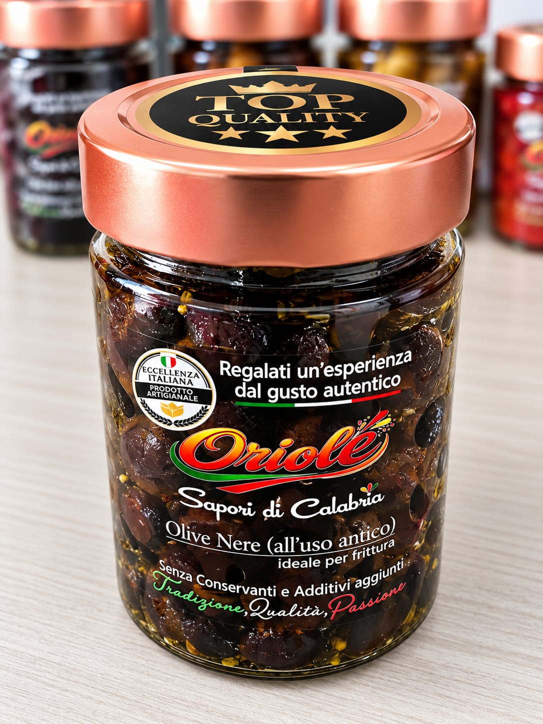 Close-up product photograph of a glass jar of dark olives with a copper-colored lid, centered against softly blurred jars and bright, even lighting.