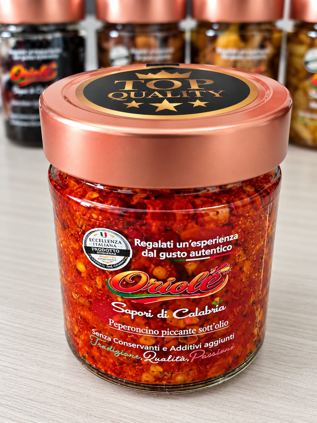 Close-up product photograph of a glass jar filled with red pepper pieces, centered on a light tabletop with blurred jars in the background under bright lighting.